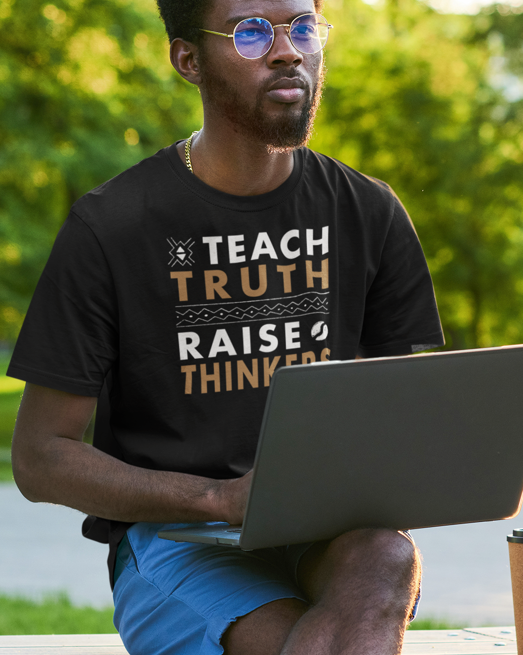 Man sitting outdoors with a laptop and coffee, wearing a t-shirt with text that says Teach Truth Raise Thinkers